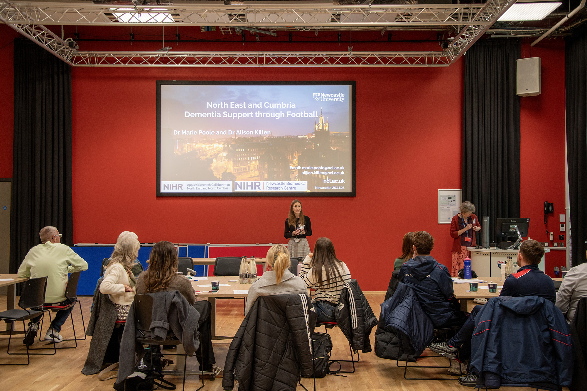 A group of people are sitting in front of a big screen, all looking to the front. In front of the screen is a woman standing to present to the group.